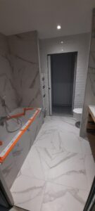 Salle de bain moderne en cours de finition, carrelage marbre blanc veiné au sol et murs, baignoire protégée par ruban orange FAP, porte coulissante.