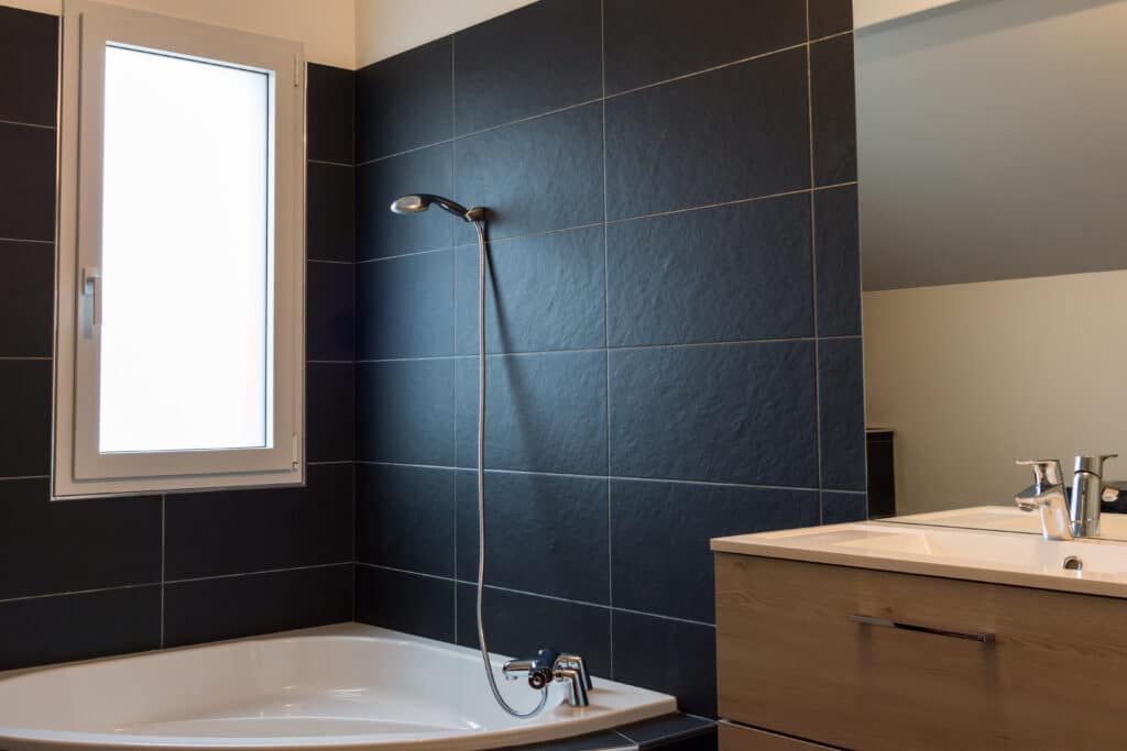 Salle de Bain Moderne: Baignoire, Carrelage Noir et Bois Salle de bain moderne avec grande baignoire blanche, mur de carreaux noirs texturés, fenêtre lumineuse et meuble-lavabo en bois.