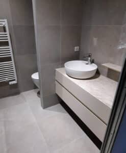 Salle de bain moderne avec vasque à poser blanche, meuble suspendu clair, carrelage gris taupe au sol et mural, et sèche-serviettes blanc.