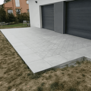 Grande terrasse en dalles de grès cérame gris clair à motifs, juxtaposée à une façade moderne blanche et portes volets roulants gris foncé.