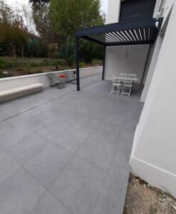 Terrasse carrelée de grandes dalles grises sous une pergola moderne noire, avec mobilier de jardin et vue sur la végétation.